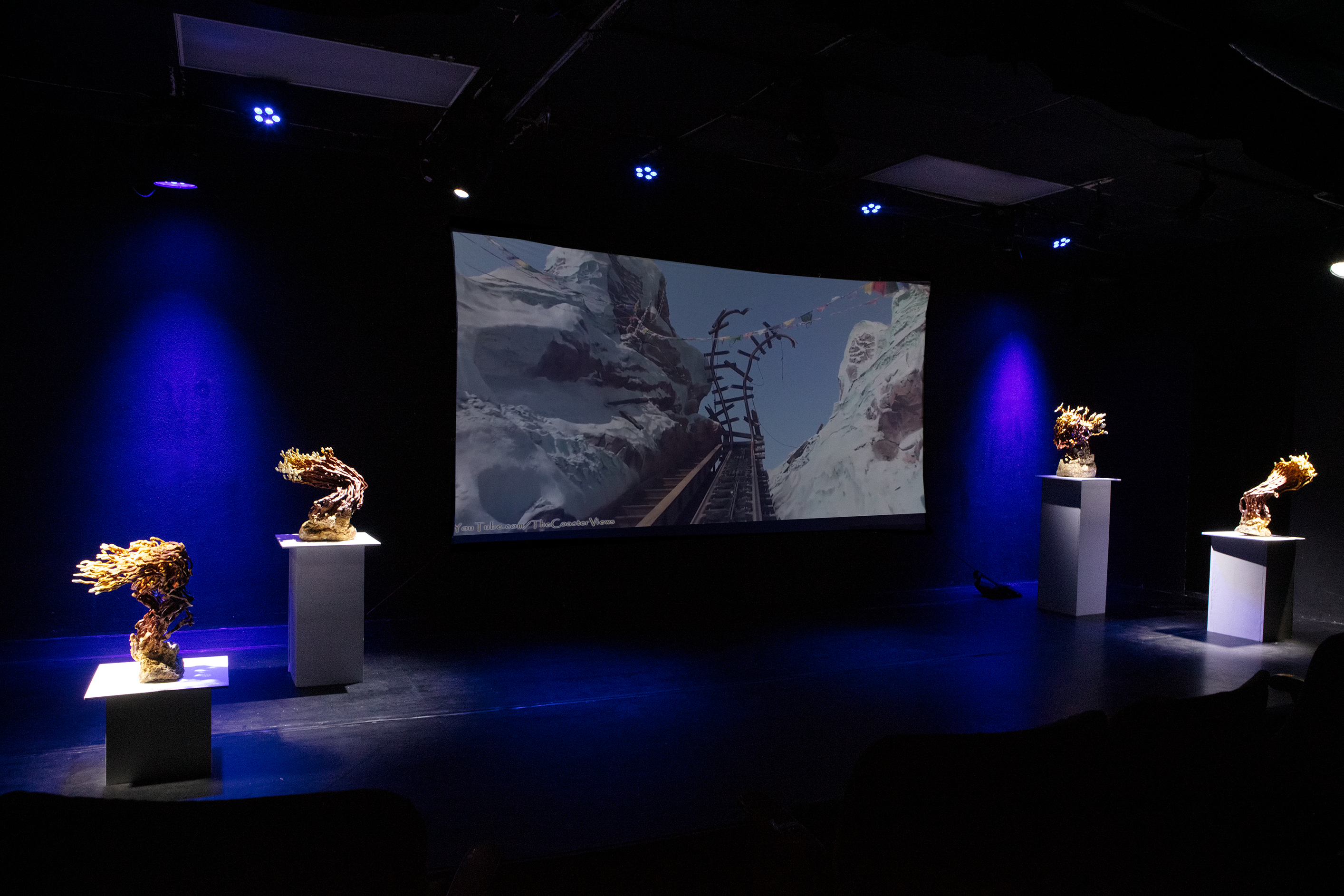 3/4 Left installaion view of a projecion screen in a dark theater with two sculptures on both the left and right sides of the screen illumated by spotlights